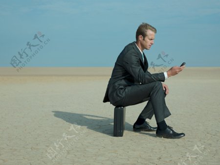 在沙漠中坐着皮箱玩手机的外国商业男人图片